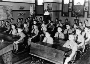 Photo showing a classroom of neatly-dressed boys seated, facing the front of the room. A nun is standing at the back.