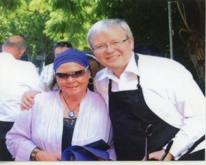 Kevin Rudd, wearing an apron and holding tongs, stands to the right, with his arm around a woman's sholders.