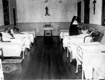 Photo showing a large room with two rows of beds. A nun is tending to the occupant of one bed.