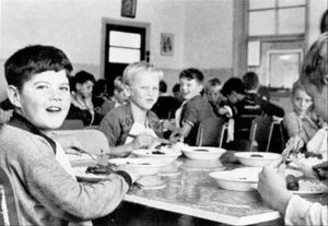 Photo showing a large room filled with smiling young boys seated at tables, with food in front of them